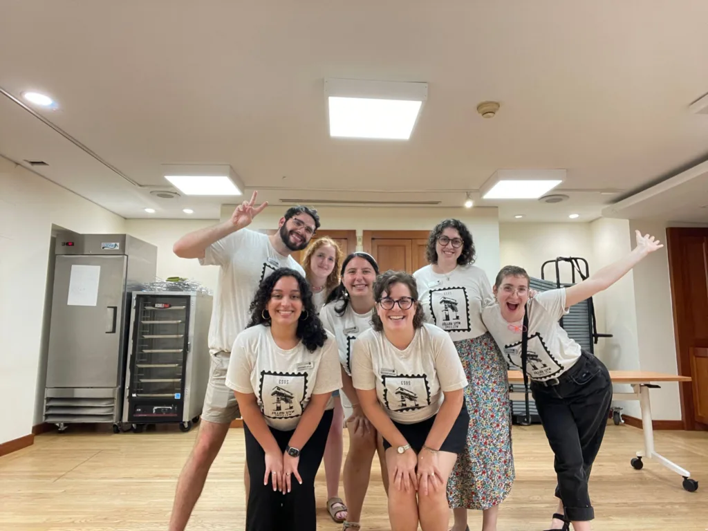 Welcome to NYU Hillel! Seven Hillel staff members wearing white and black shirts pose in an empty kitchen with big smiles and outstretched hands.