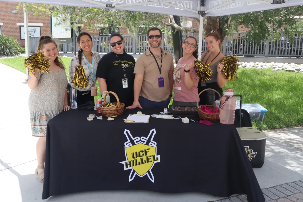 Welcome to UCF Hillel! Six Hillel staff members pose behind a table with a black UCF Hillel tablecloth on it. They are holding pompoms and smiling at the camera.